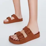 Sandalias Melissa Cozy Slide M-Lover color marrón, con diseño de dos tiras anchas con hebillas ajustables y tiras de tela con la marca estampada. Confeccionadas en PVC extra flexible, con plantilla anatómica y suela dentada.