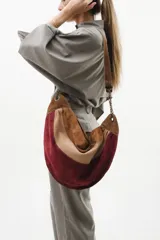 Cartera bandolera de diseño semicircular confeccionada en gamuza con combinación de colores en tonos bordó, beige, tostado y chocolate. Presenta una correa con detalle de cadena metálica en tono oro viejo y herrajes a juego. El interior se encuentra forrado en tela estampada y cuenta con bolsillos internos.