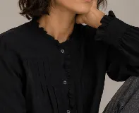 Camisa negra de manga larga con detalles de volados en el cuello alto, a lo largo de la botonadura frontal y en los puños elásticos. Presenta frunces en el canesú delantero y cierre frontal con botones.