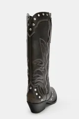 Bota estilo cowboy de caña alta, confeccionada en cuero con acabado desgastado. Presenta detalles de bordados en pespunte a contraste, aplicación de tachas metálicas plateadas y cierre frontal con cremallera. Cuenta con tacón bloque y puntera fina.
