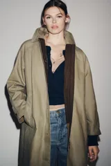 Trench coat de corte largo y diseño minimalista, confeccionado en tejido encerado color beige. Presenta cuello solapa, cierre frontal con botones oscuros y bolsillos laterales integrados.
