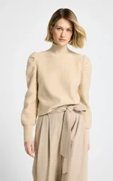 Polera de punto color beige con sutiles destellos brillantes, cuello alto y mangas largas con hombros abullonados y puños ajustados.