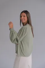 Blusa de color verde agua, con cuello en V, cierre frontal con botones y mangas largas abullonadas con puños elásticos. Presenta detalles bordados sutiles en la parte frontal y en las mangas.