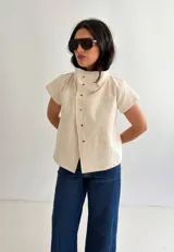 Top de algodón color crema, de manga corta y cuello alto estructurado. Cuenta con cierre frontal cruzado con botones metálicos a presión y costuras decorativas.