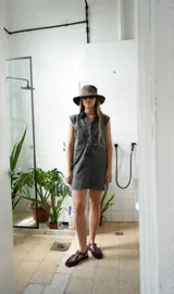 Vestido corto de jean color gris oscuro con efecto acid wash, sin mangas, con cuello camisero y cierre frontal de botones. Incluye dos bolsillos en el pecho con solapa y pespuntes decorativos.