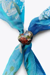 Pañuelo o bandana de seda y algodón, color fucsia intenso, con estampado floral y paisley en tonos beige/crema. Se anuda al cuello con un broche metálico en forma de corazón dorado y verde.