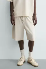 Bermuda de corte relajado, color crema o blanco roto, con textura acanalada (canalé). Presenta cintura elástica ajustable con cordón, bolsillos de vivo frontales y un bolsillo plastrón trasero.