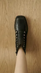 Bota corta de cuero negro con diseño versátil que incluye una pieza frontal removible con cierre y detalles de tachas metálicas. Presenta una combinación de texturas en cuero liso y cuero con textura tipo reptil en el talón, con suela track de goma.