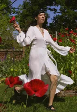 Vestido tipo batón de crepe color blanco, con escote en V, mangas largas abullonadas con puños elásticos y diseño envolvente con lazo de ajuste en la cintura. Presenta un largo irregular con volado en el ruedo.