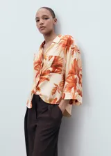 Camisa de corte recto y tejido fluido con estampado de palmeras en tonos naranja sobre fondo crema. Presenta cuello camisero, cierre frontal de botones ocultos, bolsillo en el pecho y mangas tres cuartos con vuelta.