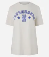 Remera de algodón color beige claro, manga corta y escote redondo, con estampado frontal en azul que dice "Copenhagen 1996" en tipografía universitaria rodeado de estrellas.