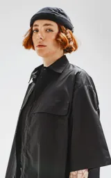 Camisa de manga corta color negro, confeccionada en popelín de algodón, con silueta oversize y dos bolsillos frontales, uno con solapa y otro con ojal y botón.