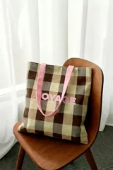 Tote bag de tela con diseño a cuadros en tonos marrón, beige y verde, tiras de hombro color rosado y la palabra "VOYAGE" estampada en el frente en color rosado.