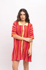 Vestido camisola corto de tejido fresco de algodón, con diseño a rayas verticales en tonos rojos, naranjas y fucsias. Cuenta con cuello polo y botonadura frontal en tono amarillo claro, y mangas cortas holgadas.