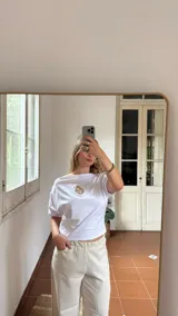 Remera blanca de algodón con cuello tipo bote y detalle de elástico en los hombros, creando un efecto de caída asimétrica.