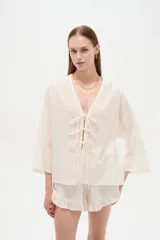 Blusa de algodón blanco con diseño de broderie, cuello en V y cierre con lazos frontales, mangas largas de corte holgado.