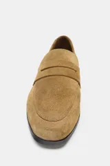 Mocasín plano de mujer en serraje color camel, con antifaz y copete. Presenta punta redonda y suela baja de goma negra.
