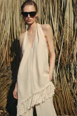 Top halter color crudo, de escote en V y diseño confeccionado en hilatura con mezcla de viscosa. Presenta bajo asimétrico con flecos al tono y cierre en la espalda.