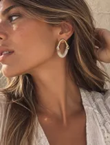 Aros colgantes con diseño geométrico ovalado, combinando una estructura superior dorada con una pieza inferior de acrílico translúcido blanco.