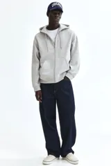 Campera con capucha de felpa gris melange, con cierre frontal, bolsillos tipo canguro y cordón ajustable. Presenta hombros caídos, corte holgado y puños y bajo en punto acanalado.