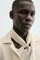 Camisa de calce relajado confeccionada en tejido con algodón de color beige. Presenta cuello bowling, manga corta, aberturas laterales en el bajo y cierre frontal con botones.