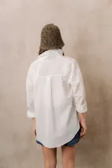 Camisa blanca de calce amplio, confeccionada en tejido liviano con sutiles rayas verticales, cuello clásico, mangas largas con hombros caídos y abotonadura frontal.