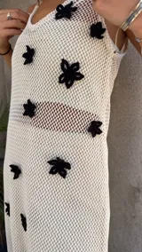 Vestido largo de tejido de red blanco, con apliques de flores negras en relieve. Lleva tirantes finos y tiene un corte recto con abertura lateral. Se usa sobre una prenda interior blanca.