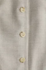Chaleco largo de vestir color beige claro, con cuello en V, cierre frontal de botones y bolsillos ribeteados delanteros. Presenta un corte entallado y forro interior.
