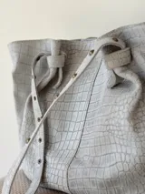 Cartera tipo shopper de cuero gris con textura símil cocodrilo, diseño amplio, correas ajustables con tachas y cierre superior.