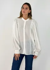 Camisa blanca de corte recto y diseño fluido, con cuello mao, cierre frontal de botones y mangas largas abullonadas con detalle de frunces en los hombros y puños abotonados.