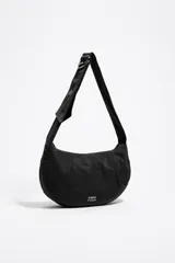 Bolso tipo media luna (hobo bag) de nylon negro, con costuras verticales que le dan estructura y un asa de hombro ajustable con hebilla metálica. Presenta el logo de Bimba y Lola en blanco en la parte inferior central.