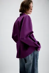 Camisa de manga larga en color violeta, con cuello solapa y cierre frontal de botones. Presenta un diseño de corte recto con detalle de pliegues en la espalda.