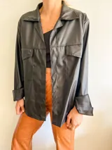 Camisa negra de eco cuero con cuello clásico, solapas y cierre frontal con botones a presión. Tiene dos bolsillos de parche en el pecho y mangas largas con puños abotonados.