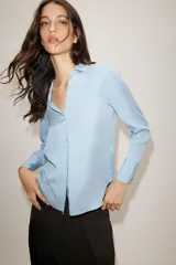 Camisa de cuello solapa y escote en pico, con manga larga y cierre frontal mediante botones. Presenta un diseño fluido con bajo asimétrico y aberturas laterales.