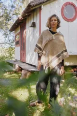 Poncho artesanal de lana en color beige, con diseño de tejido rústico y detalles de guardas geométricas en negro y blanco en los bordes. Presenta terminaciones con flecos largos en todo el contorno.