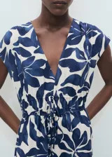 Vestido largo de corte evasé con estampado botánico en azul sobre fondo crudo. Presenta cuello de pico, mangas cortas y un panel fruncido en la cintura con cordón ajustable.