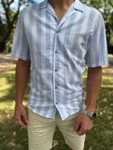 Camisa de manga corta con diseño a rayas verticales en color celeste y blanco, cuello abierto estilo cubano y cierre frontal con botones marrón claro.