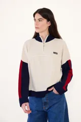 Sweater de punto acanalado con cuello alto y cierre frontal parcial. Presenta un diseño tricolor en bloques con cuerpo color crema, cuello y puños azul marino, y detalles en rojo en las mangas. Incluye una pequeña etiqueta con logo en el pecho.