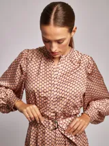Vestido midi de satén con estampado geométrico en tonos marrones y naranjas. Presenta cuello mao, mangas largas abullonadas con puños ajustados y un cinturón a tono con hebilla metálica que marca la cintura.