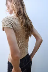 Top de punto color beige con cuello subido, manga corta y diseño texturizado con efecto arrugado. Presenta aplicaciones de perlas distribuidas en la parte frontal y las mangas.