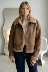 Campera tipo gamulán color camel, confeccionada en gamuza sintética con interior y detalles de corderito al tono en cuello, puños y ruedo. Presenta cierre frontal metálico y hebilla ajustable en el cuello.