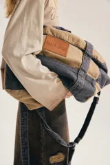 Bolso estilo duffel o bandolera de diseño bicolor, combinando lona en tono tostado y denim gris gastado. Cuenta con asas superiores, correa ajustable para el hombro y un parche de cuero marrón con el logotipo de la marca grabado en la parte frontal.