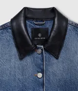 Chaqueta corta de denim azul con cuello camisero de cuero negro y cierre frontal de botones metálicos. Presenta dos bolsillos delanteros con solapa y hombros caídos.