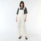 Enterito de jean color blanco con diseño de pierna ancha, bolsillo frontal en el pecho y tirantes ajustables con hebillas metálicas.