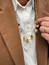 Collar de cadena fina dorada con múltiples dijes colgantes, incluyendo una moneda, una esfera brillante, una piedra verde, hojas, un dije de flores con perlas y un corazón calado.