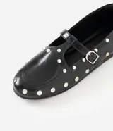 Zapato tipo salomé de color negro con acabado liso, puntera redondeada y suela plana. Presenta una tira central con hebilla metálica ajustable y está decorado con tachuelas metálicas plateadas distribuidas por toda la superficie.