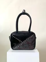 Cartera rígida negra con detalles en rojo y tachas plateadas. Tiene herrajes y cierre metálico plateado marca YKK, asa fina para llevar al hombro o en la mano y correa larga extra regulable. El interior es forrado, tiene un bolsillo con cierre enmarcado en cuero, y un segundo bolsillo plaqué.