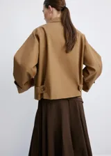 Chaqueta corta de algodón color marrón tostado, con cuello alto, cierre frontal de botones, bolsillos ribeteados y mangas largas con trabillas ajustables en los puños.
