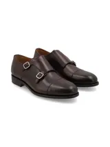 Zapato tipo Monk Strap de cuero color marrón oscuro, con doble hebilla metálica plateada y puntera reforzada.