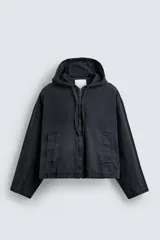 Cazadora tipo bomber de denim color negro con efecto lavado, corte boxy fit, cierre frontal de cremallera, capucha ajustable con cordón y bolsillos laterales de vivo.